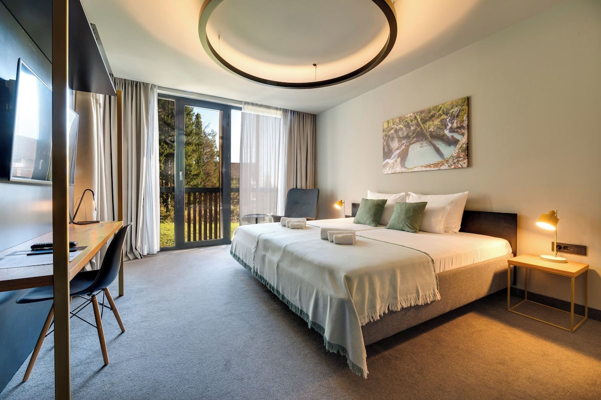 Una habitación de hotel moderna con una cama grande, un escritorio y una silla. Hay una ventana con cortinas, un cuadro en la pared y una mesita de noche con una lámpara.