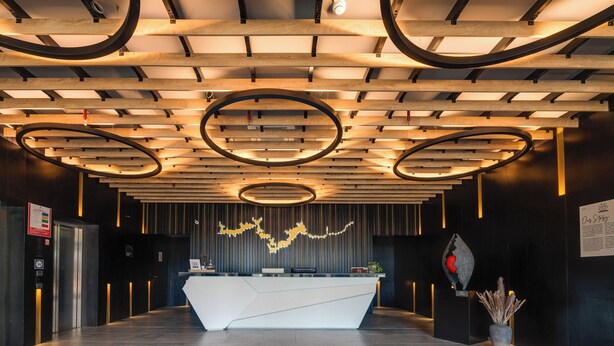A modern reception area with a white reception desk, wooden ceiling with circular light fixtures, and a black wall with a red sculpture. - Lyra Hotel Plitvice (Plitvicka Jezera)