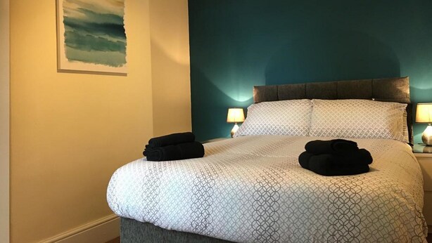 Una cama bien hecha con colcha de patrón geométrico, dos almohadas y dos toallas dobladas. Encima de la cama hay un cuadro colgado en la pared, y a la derecha, una mesita de noche con una lámpara.