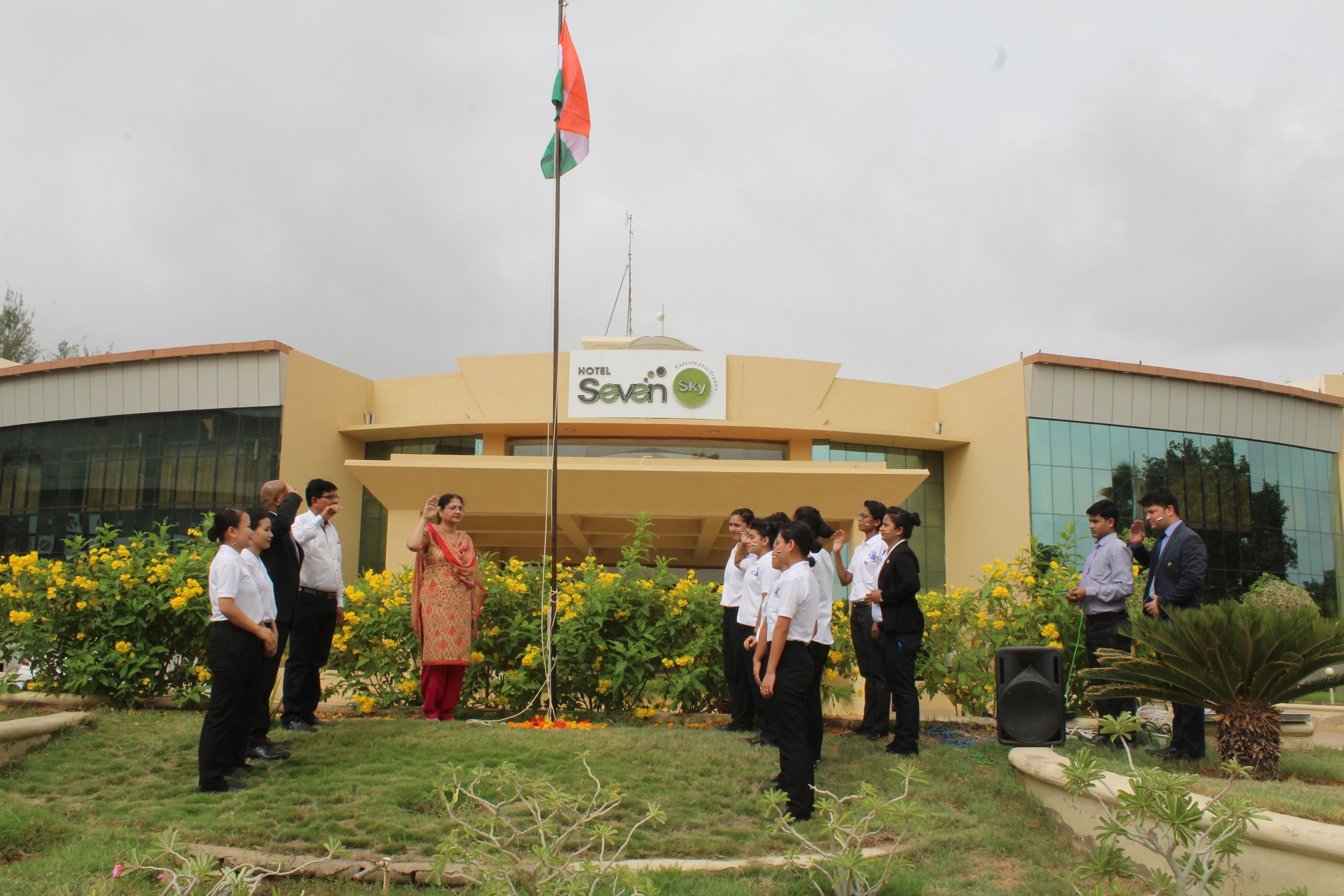A group of people standing in front of a building with a flag, a sign reading "Hotel Seven Sky", and a garden with yellow flowers.