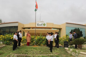 A group of people standing in front of a building with a flag, a sign reading "Hotel Seven Sky", and a garden with yellow flowers.