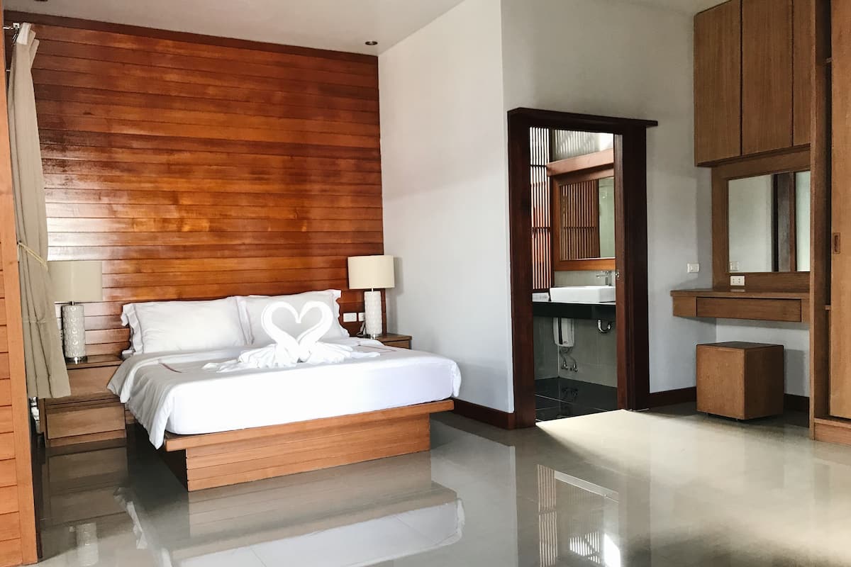 Habitación de hotel con cabecera de madera, cama con ropa blanca, mesita de noche con lámpara, baño con espejo y un pequeño armario de madera.