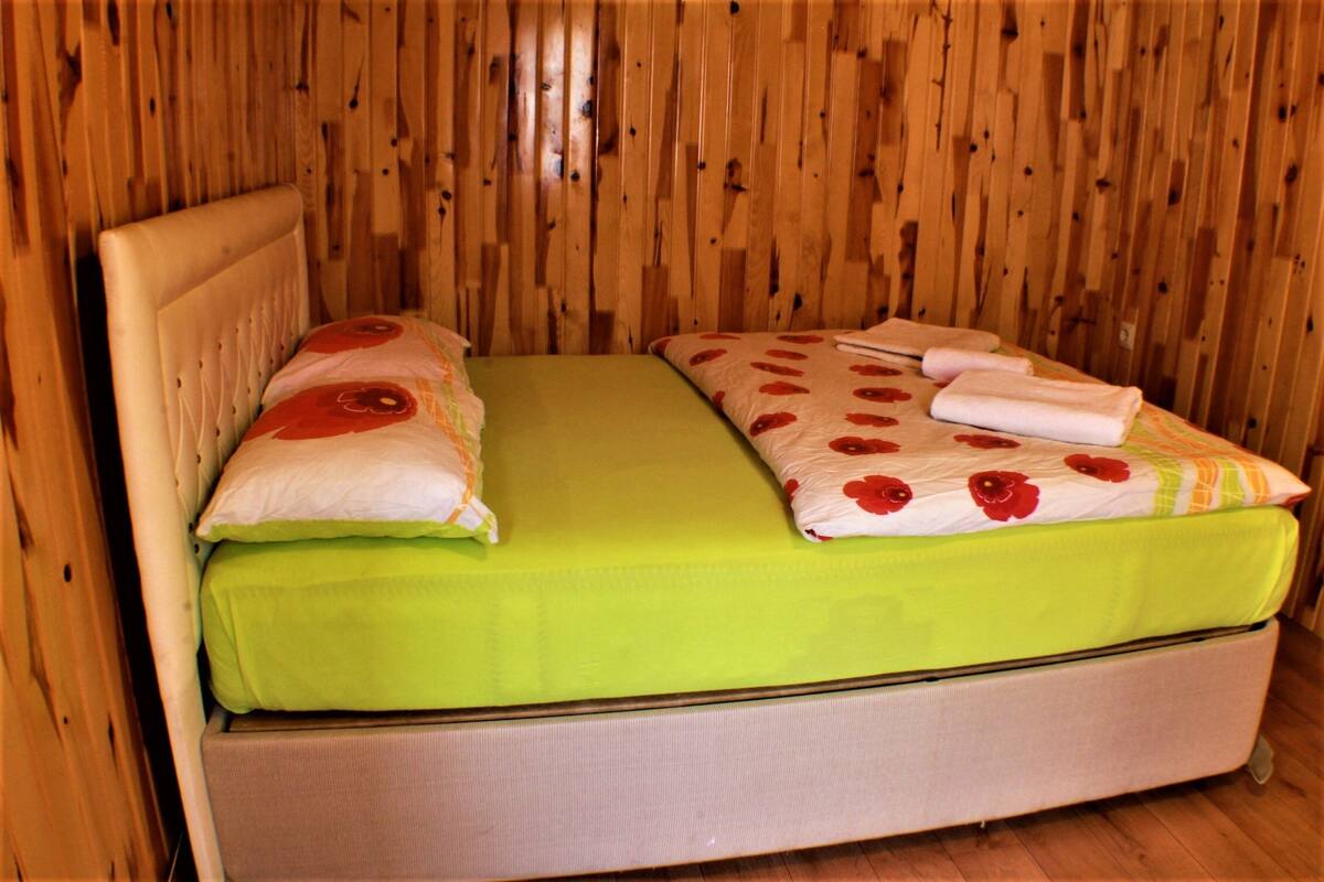 Una cama individual con colchón verde y ropa de cama estampada, frente a una pared de madera.