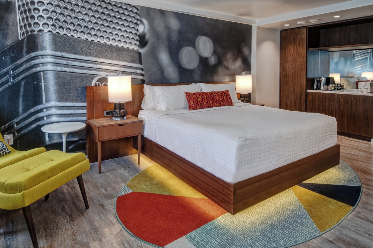 A modern hotel room with a large bed, a nightstand, a lamp, a small round table, and a yellow armchair.