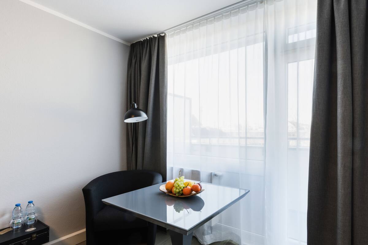 A hotel room with a glass table, a black chair, a fruit bowl, and a lamp.