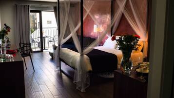 A bedroom with a four-poster bed, a bedside table with a lamp, and a view of the outside balcony.
