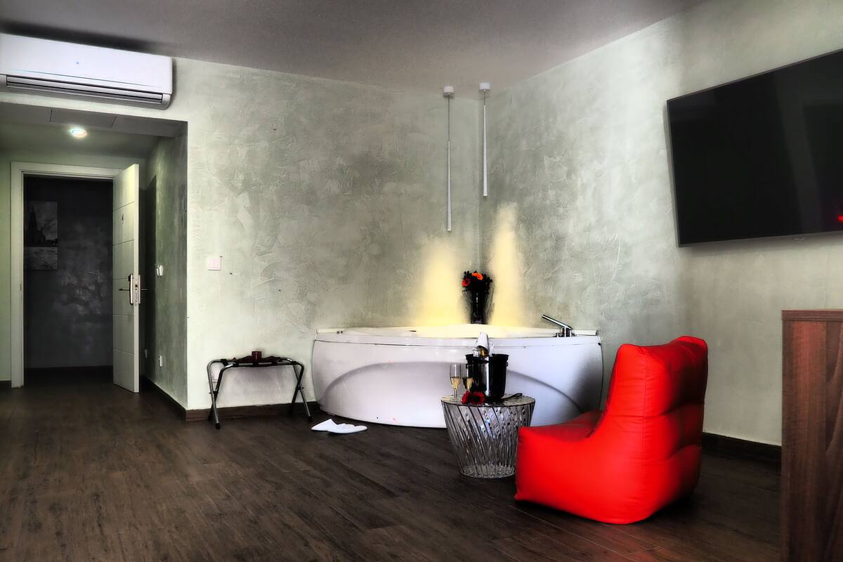 A modern bathroom with a bathtub, a red chair, a glass container with flowers, and a flat-screen TV mounted on the wall.