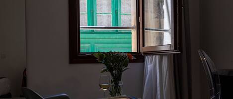 A room with a wooden floor, a rocking chair, a small table with a vase and a glass of wine, and a window with a green door visible through it.