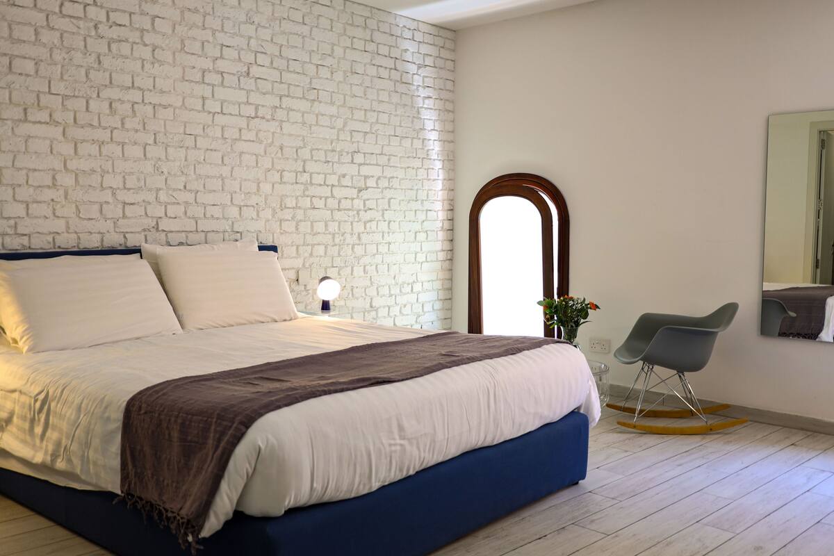 A bedroom with a white brick wall, a large bed with a blue headboard, a bedside table with a lamp, and a rocking chair.