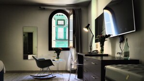 Quarto moderno com piso de madeira, uma escrivaninha com abajur, uma cadeira, uma cama e uma janela grande com cortinas.