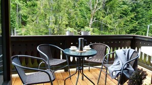 Terraza de madera con sillas de mimbre y una mesa, con vista a un bosque frondoso.