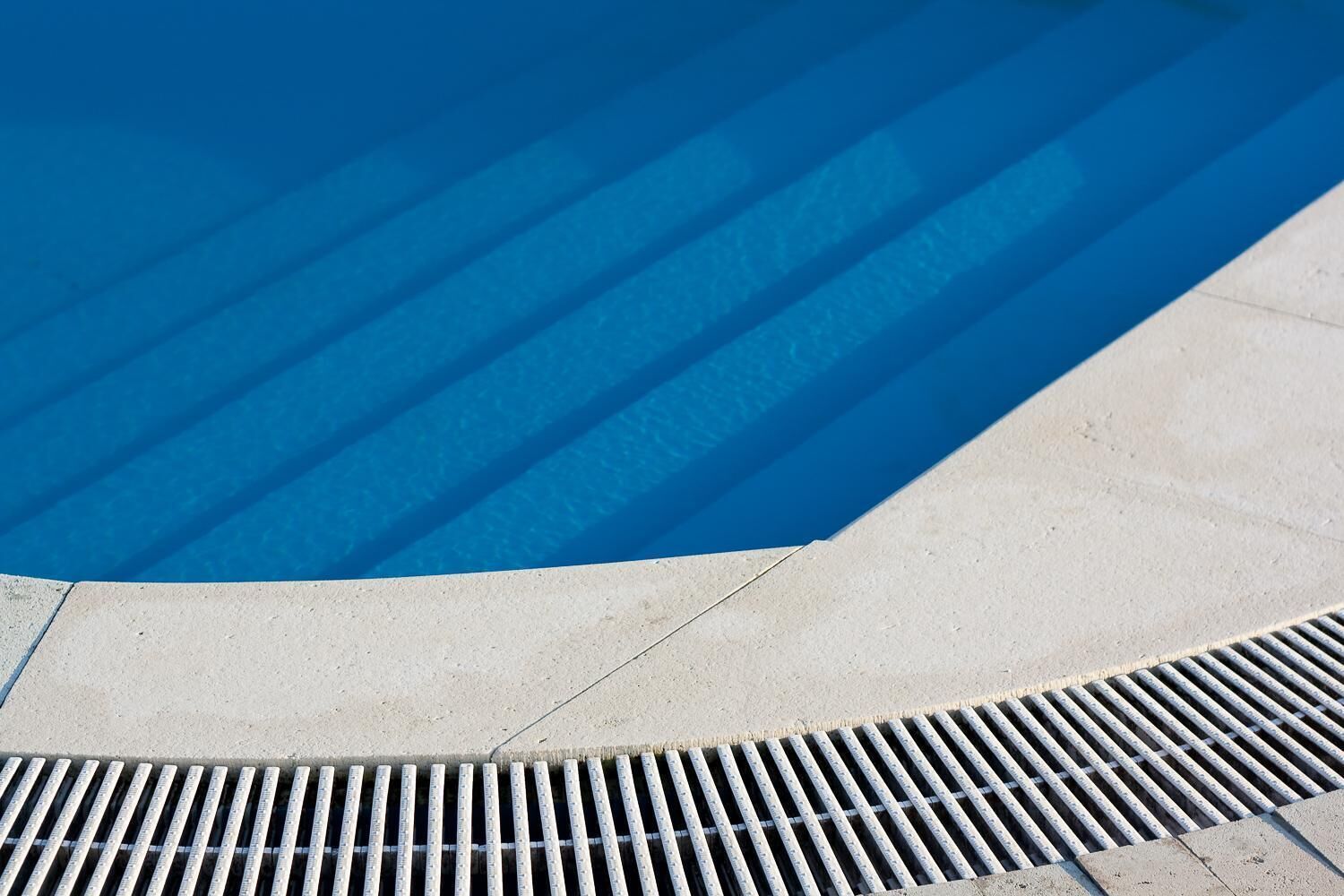 Una piscina con superficie azul cristalina, borde de concreto y una rejilla metálica en el fondo.