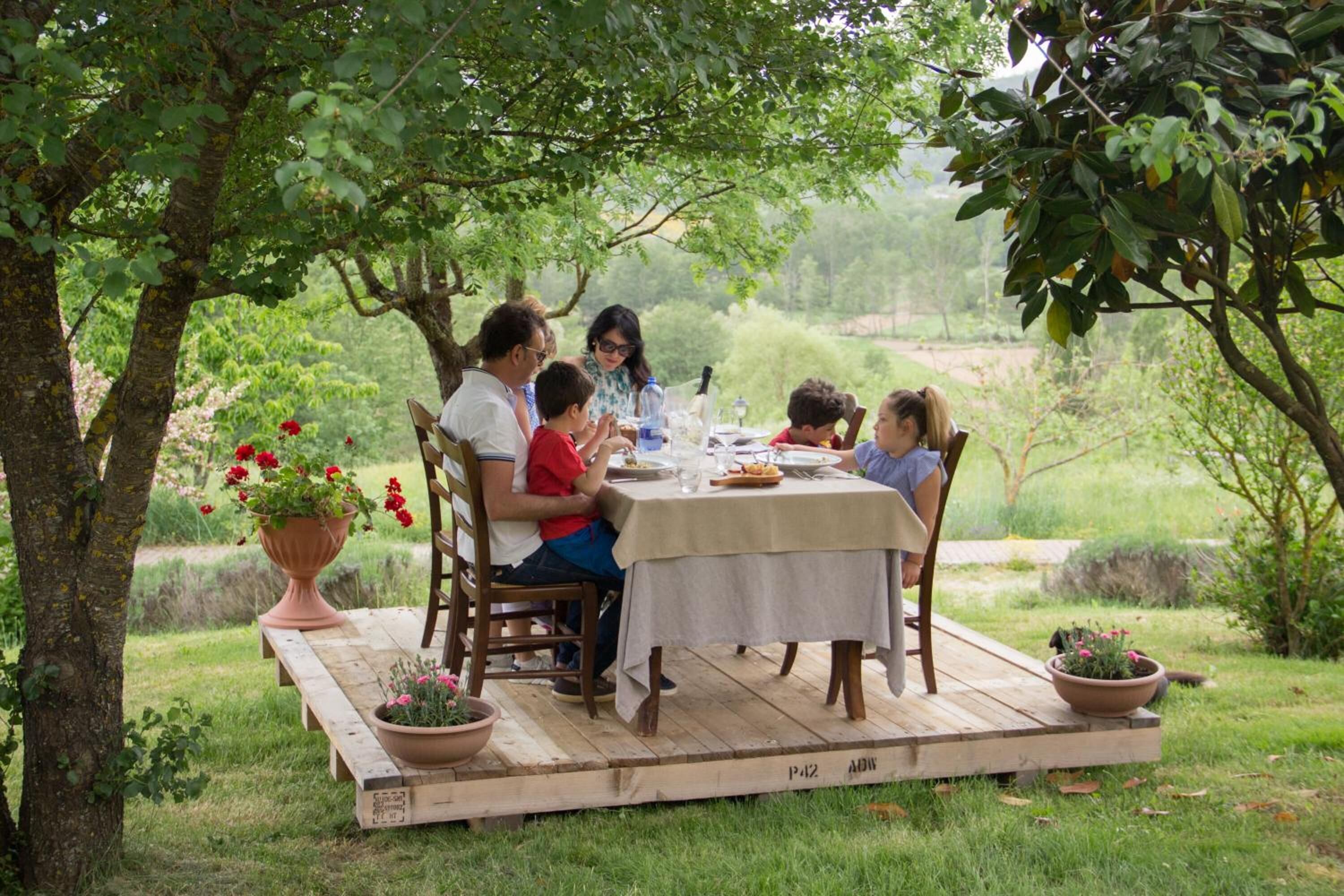 Un groupe de personnes est assis à une table en plein air, installée sur une plateforme en bois au milieu d’un jardin luxuriant.