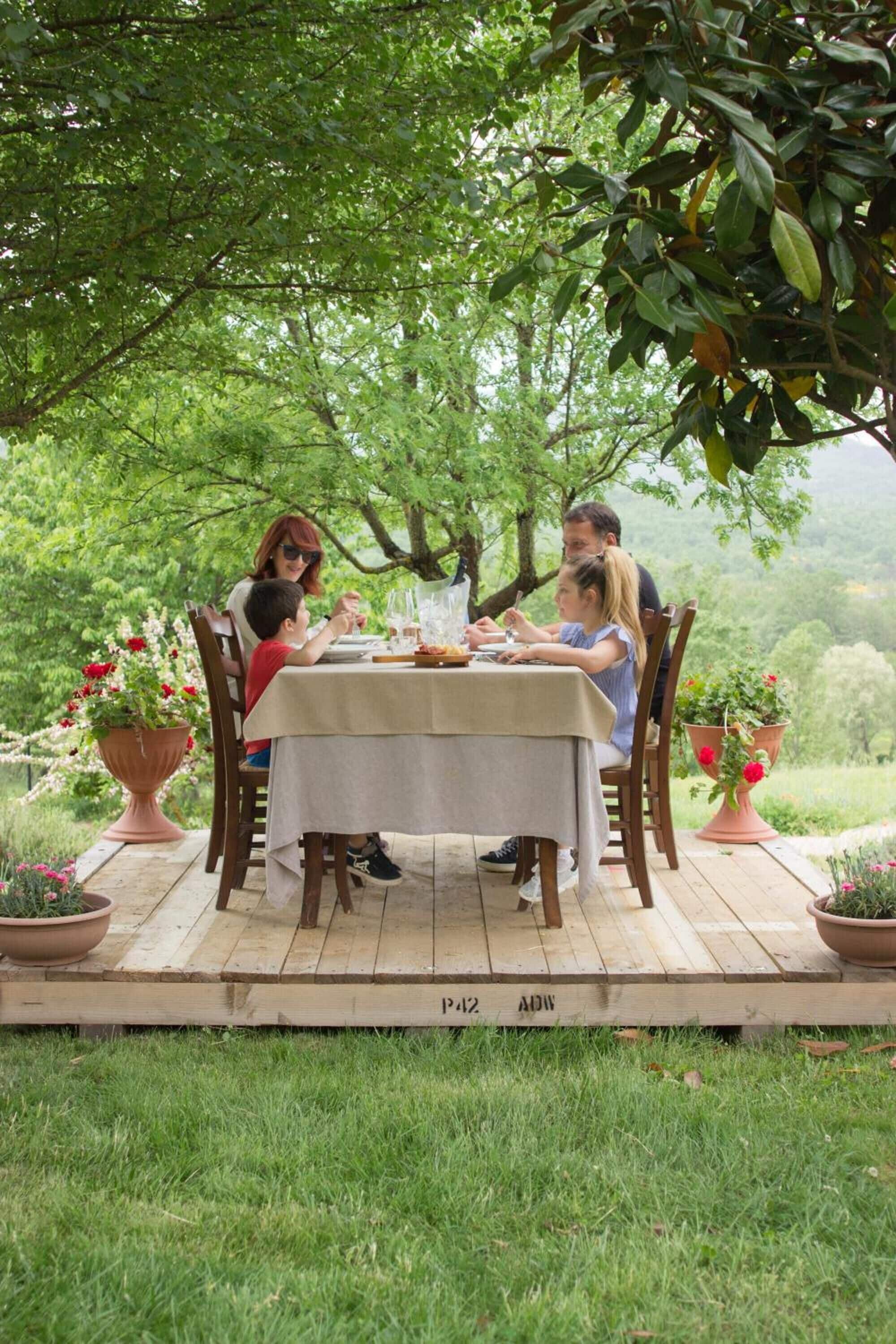 A family dining outdoors on a wooden deck with a table set for four, surrounded by lush greenery and potted plants.