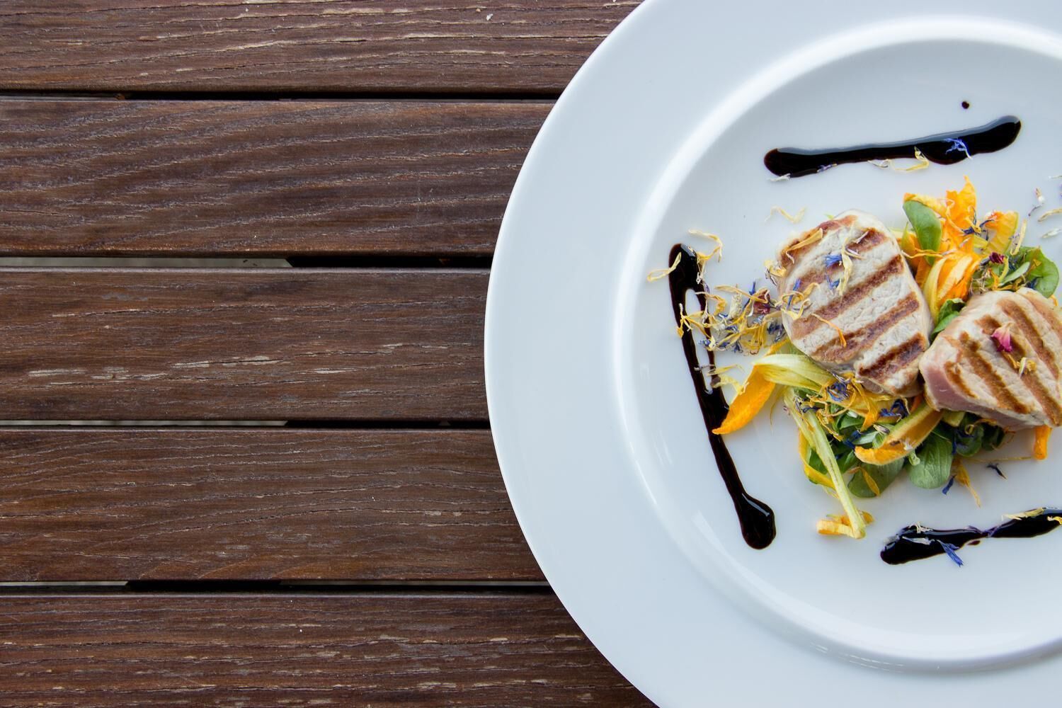 A white plate with a grilled meat dish, greens, and a dark sauce on a wooden table.