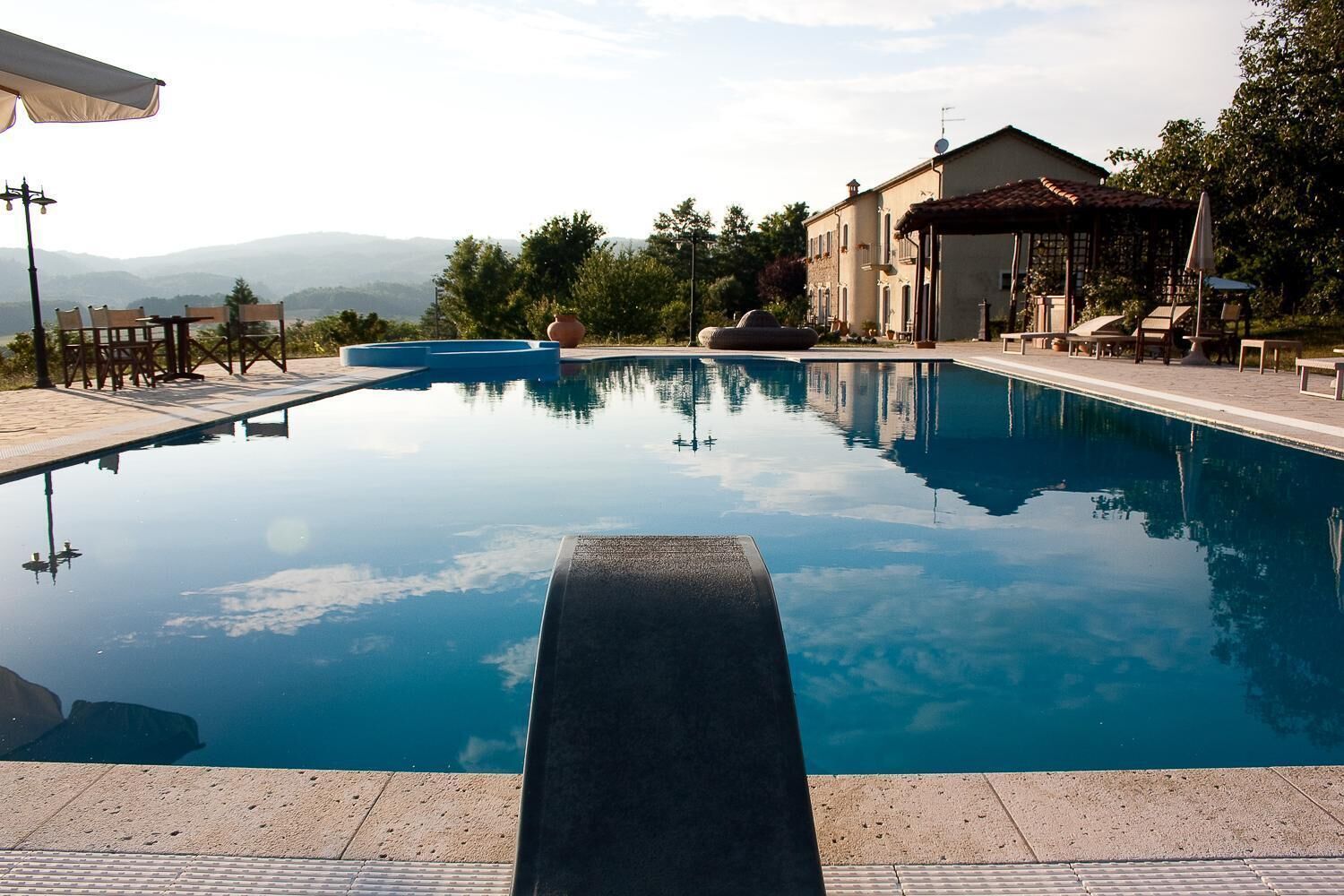 A clear swimming pool with a view of distant mountains, surrounded by outdoor seating and a building with a tiled roof.