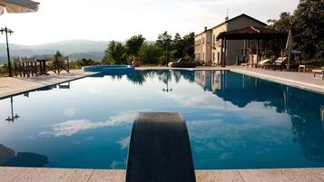 A clear swimming pool with a view of distant mountains, surrounded by outdoor seating and a building with a tiled roof.
