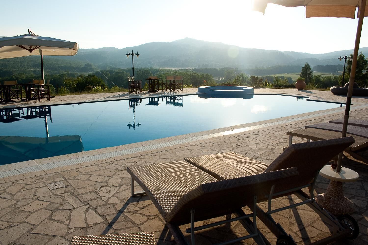 Una piscina all'aperto con lettini e tavoli, circondata da un patio in pietra e vista su montagne lontane.