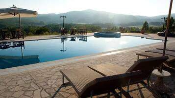 Una piscina all'aperto con lettini e tavoli, circondata da un patio in pietra e vista su montagne lontane.