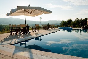 Zona de piscina con vistas a las montañas, mobiliario exterior y una sombrilla grande.