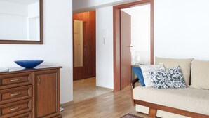 Een kamer met een houten kast, een blauwe kom erop, een beige bank met gekleurde kussens en een houten deur die naar een andere kamer leidt.