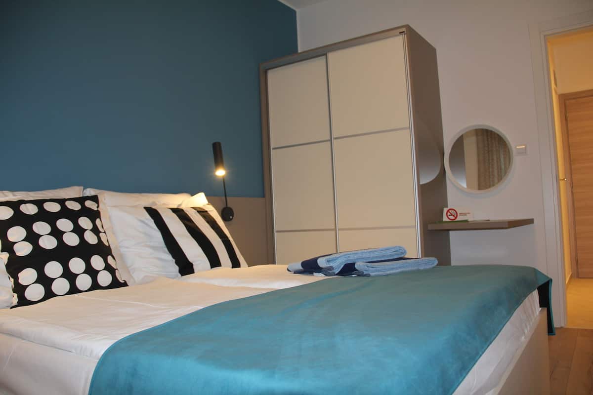 A neatly made bed with a teal blanket, a polka dot pillow, and striped pillows. There is a wardrobe with a mirror above it and a small shelf with towels.