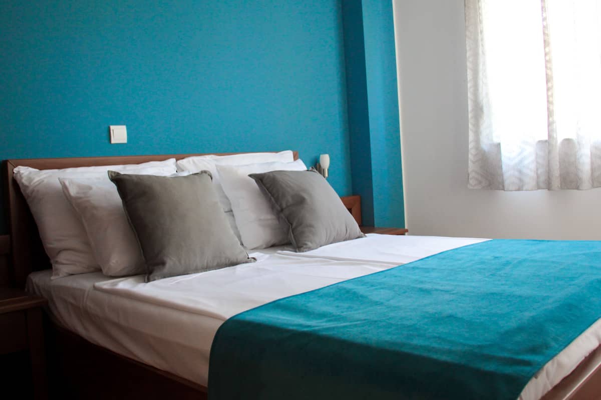 A neatly made bed with a teal bedspread and multiple pillows, a wooden headboard, and a window with sheer curtains.