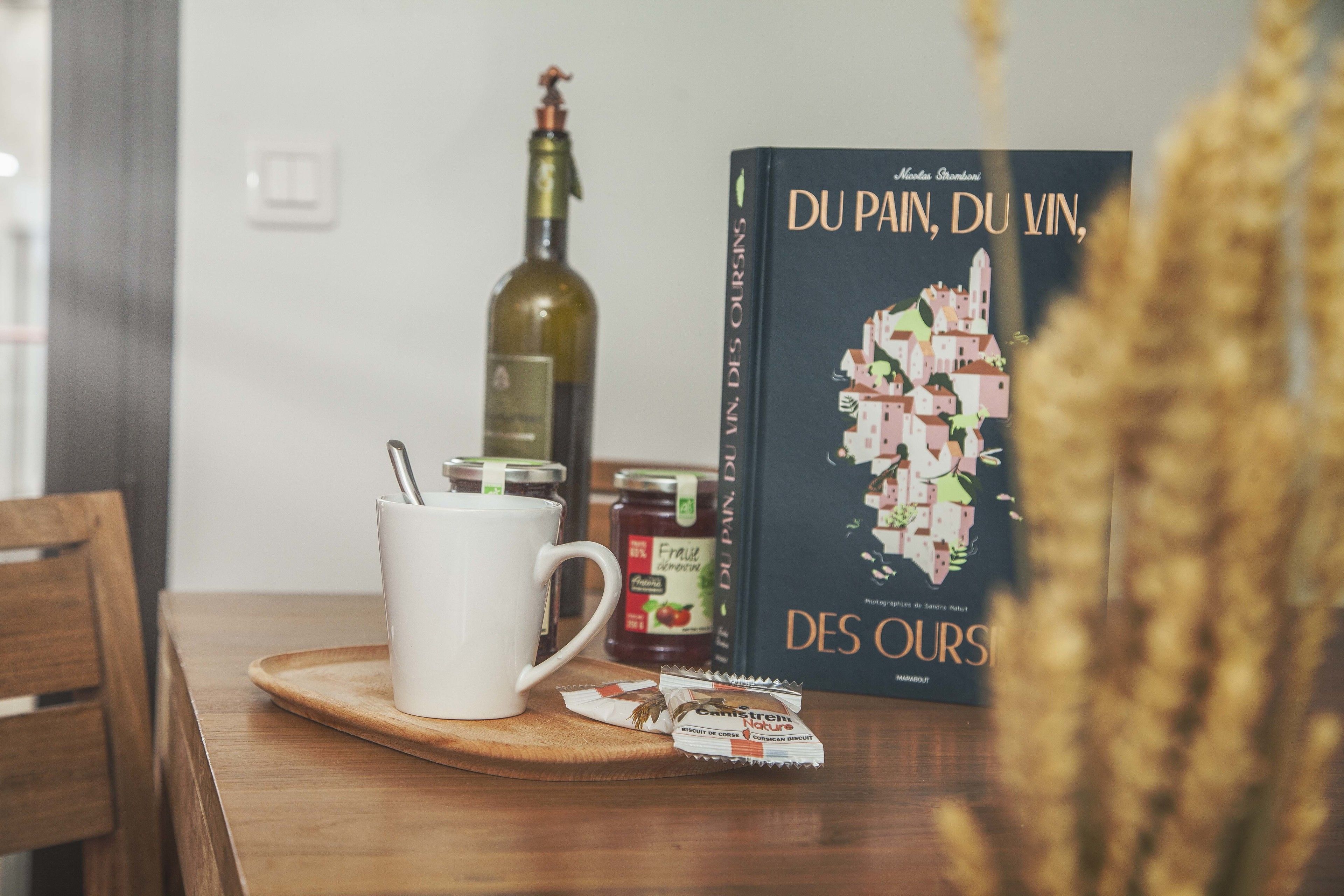 A table with a white mug, a bottle of wine, a book titled 'DU PAI, DU VIN, DES OURS', and a packet of biscuits.