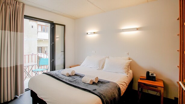 A hotel room with a bed, a bedside table with a phone, a window with curtains, and a balcony with a chair.
