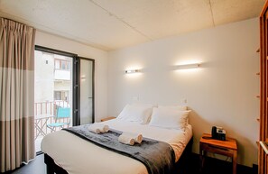 Ein Hotelzimmer mit Bett, Nachttisch mit Telefon, Fenster mit Vorhängen und Balkon mit Stuhl.