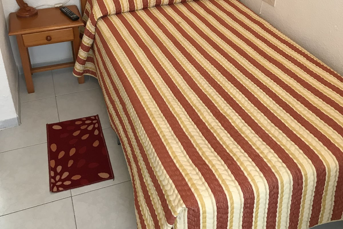 A bed with a striped bedspread, a wooden nightstand with a lamp, and a red and beige rug.