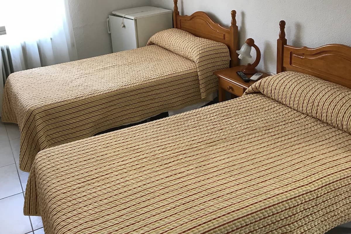 A hotel room with two single beds, a small table with a lamp, and a radiator.