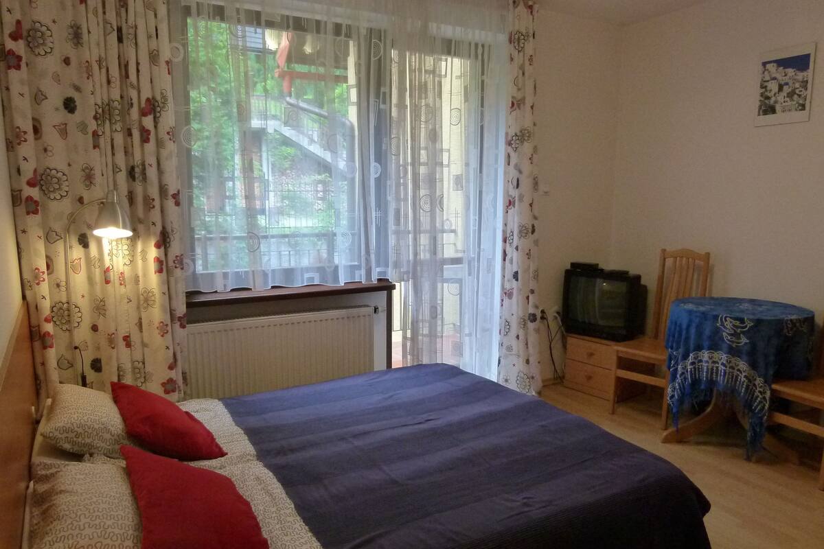 Een slaapkamer met een bed, een radiator, een televisie, een tafel met tafelkleed en gordijnen.