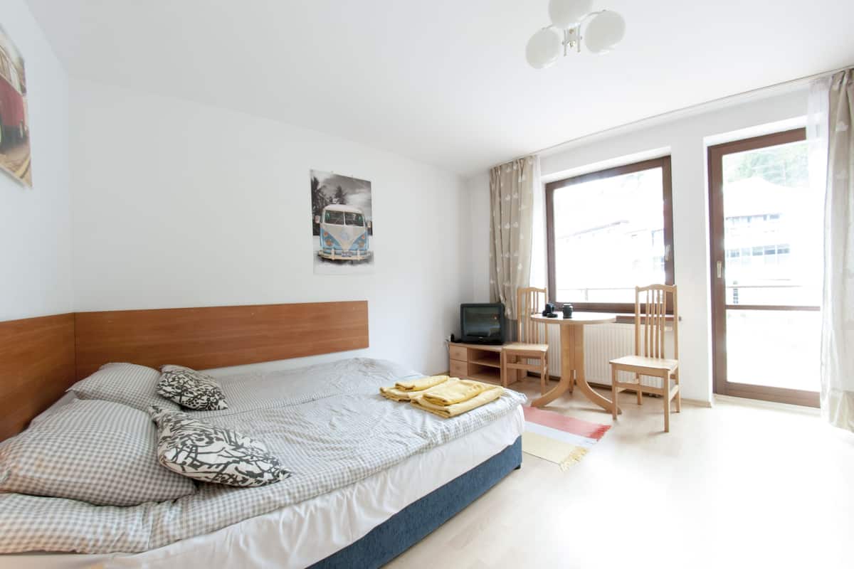 Een slaapkamer met een bed, een bureau, een stoel, een televisie en een raam met gordijnen.