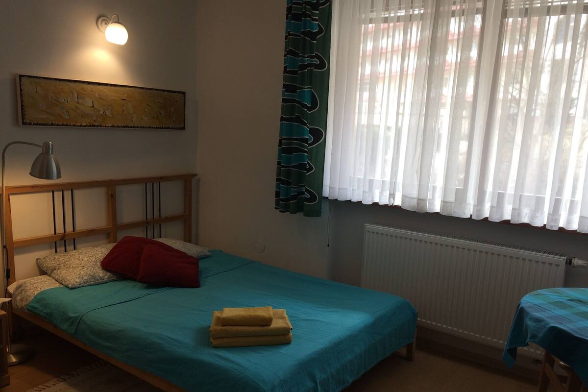 Een enkel bed met een blauwgroene sprei, een rood kussen en een opgevouwen handdoek. Er is een houten hoofdbord, een wandlamp en een radiator onder het raam.