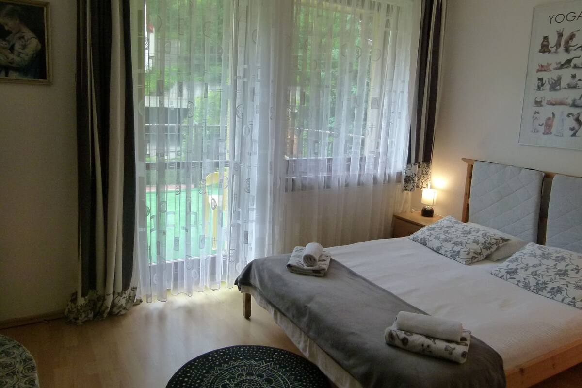 Een net opgemaakt bed met een kussen en een opgerolde handdoek, een nachtkastje met een lamp, en een raam met dunne gordijnen.