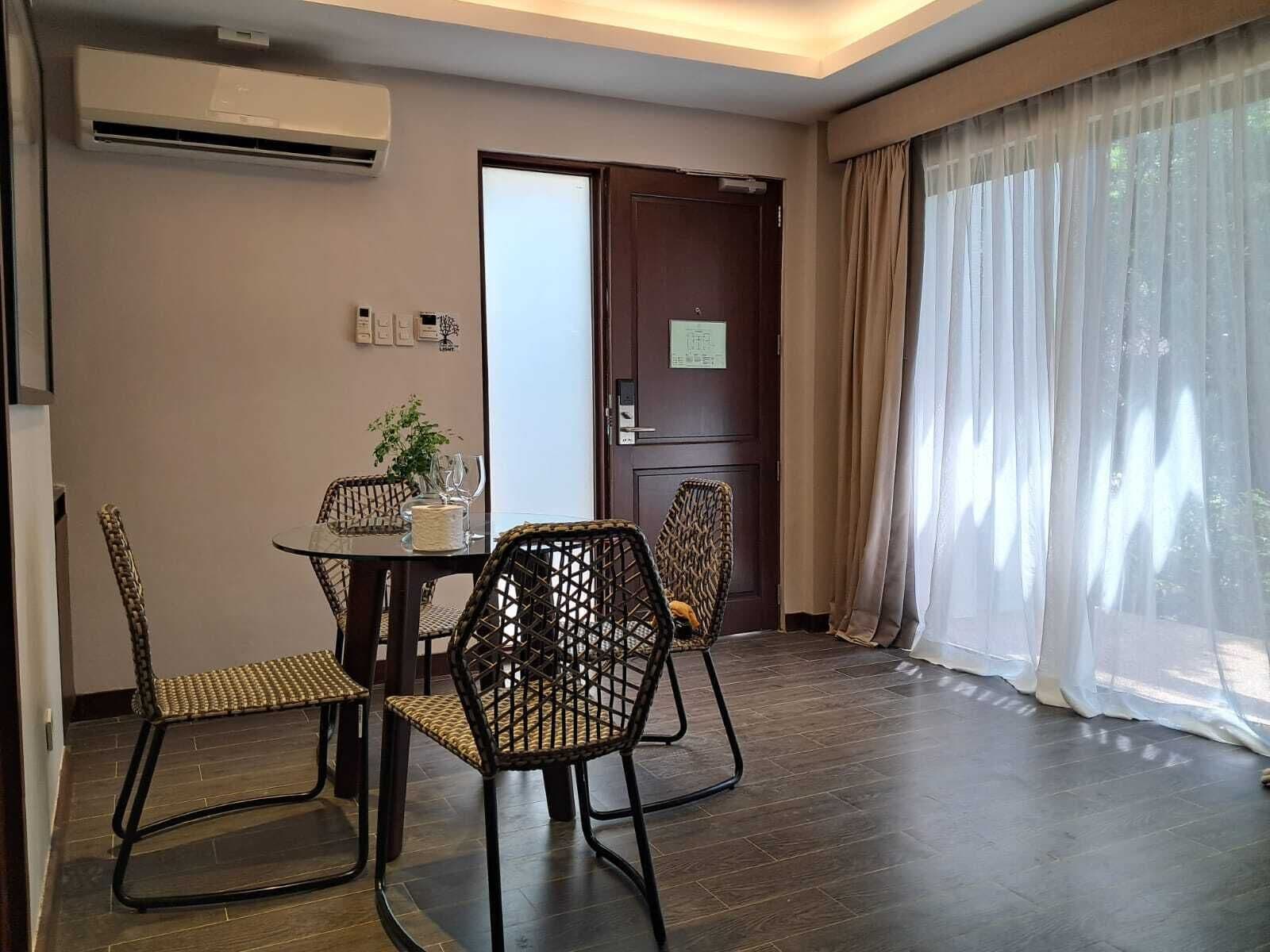 A hotel room with a dining table, four wicker chairs, a potted plant, and a large window with sheer curtains.