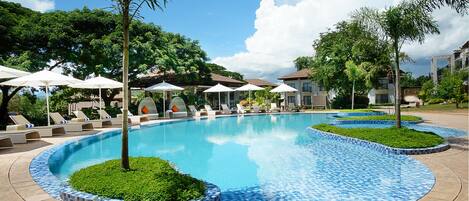 A swimming pool with lounge chairs and umbrellas, surrounded by greenery and buildings.