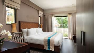 Quarto de hotel com uma cama grande, cabeceira de madeira, cadeira de vime, uma pequena mesa e vista para o verde pela janela.