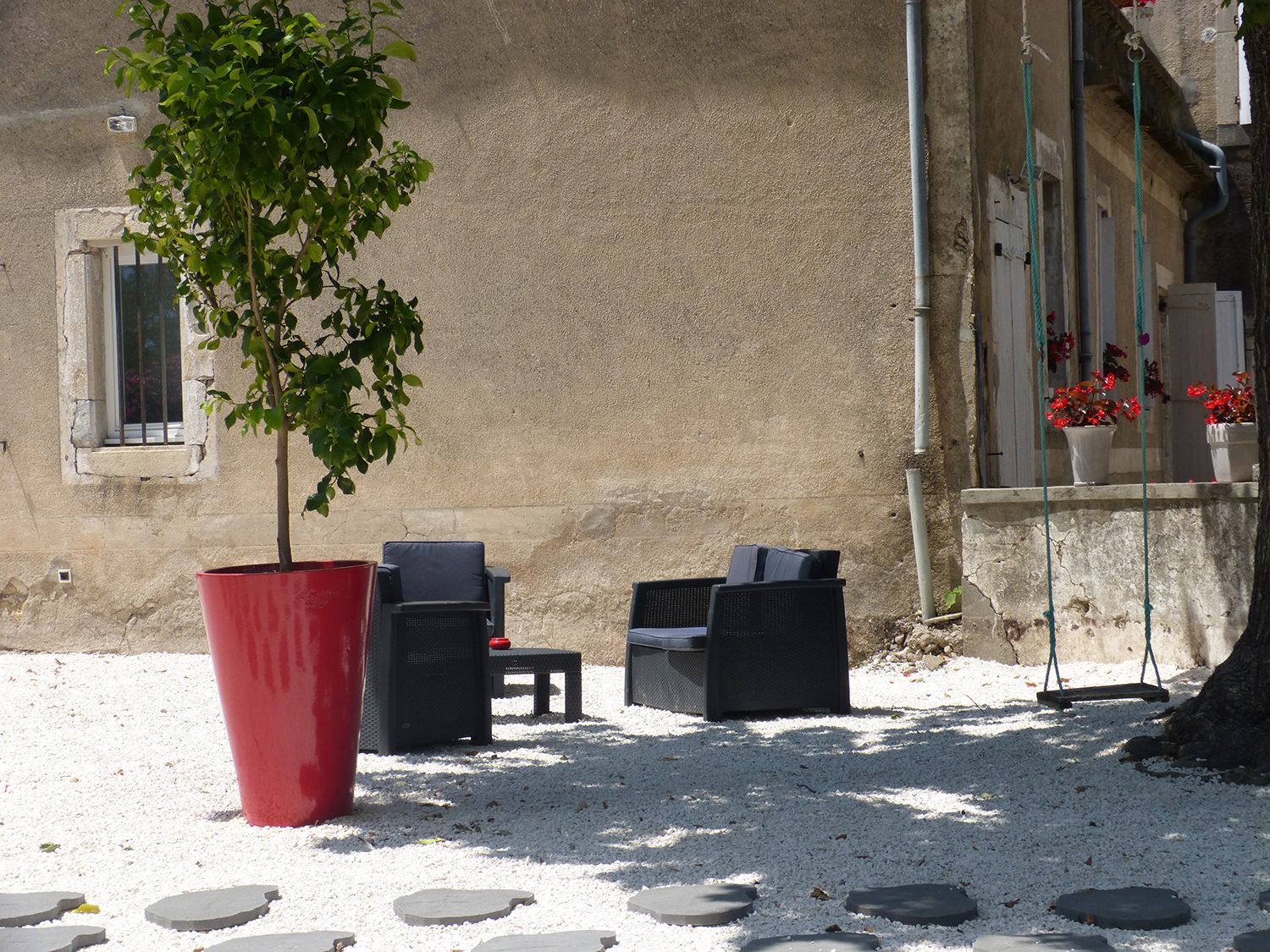 Un espace extérieur aménagé avec deux chaises et une table, un grand pot rouge contenant un arbre, et un bâtiment avec un mur beige et une fenêtre.