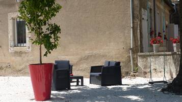 Un espace extérieur aménagé avec deux chaises et une table, un grand pot rouge contenant un arbre, et un bâtiment avec un mur beige et une fenêtre.
