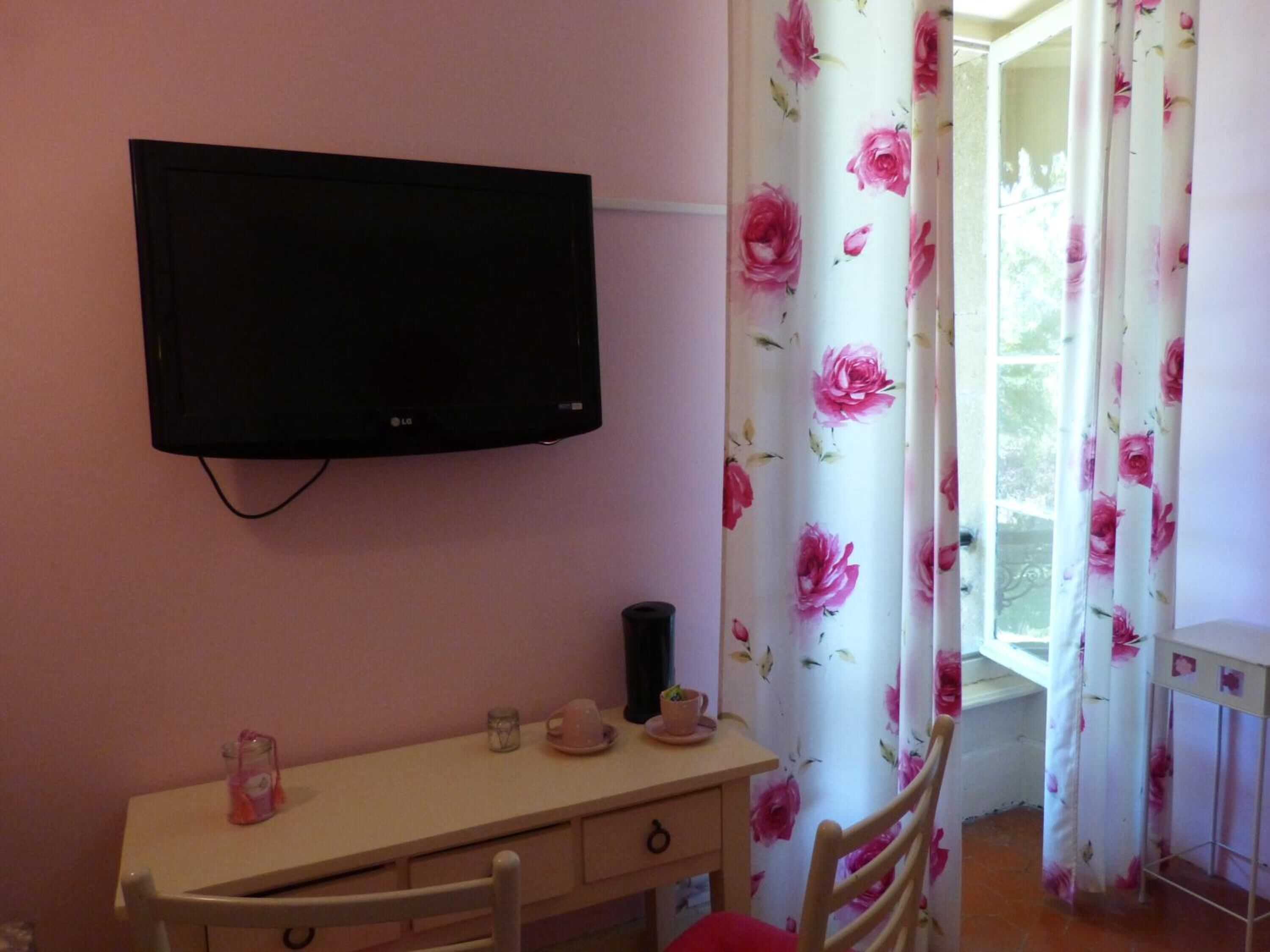 A room with a TV mounted on the wall, a desk with a chair, a small table with a cup and saucer, and floral curtains.