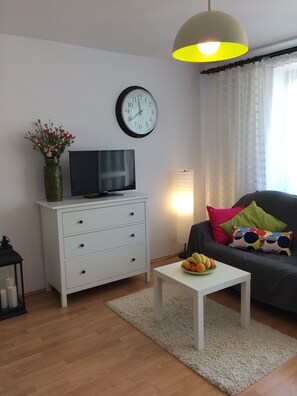 A modern living room with a white TV stand, a clock, a vase with flowers, a sofa with colorful cushions, and a small table with fruit.