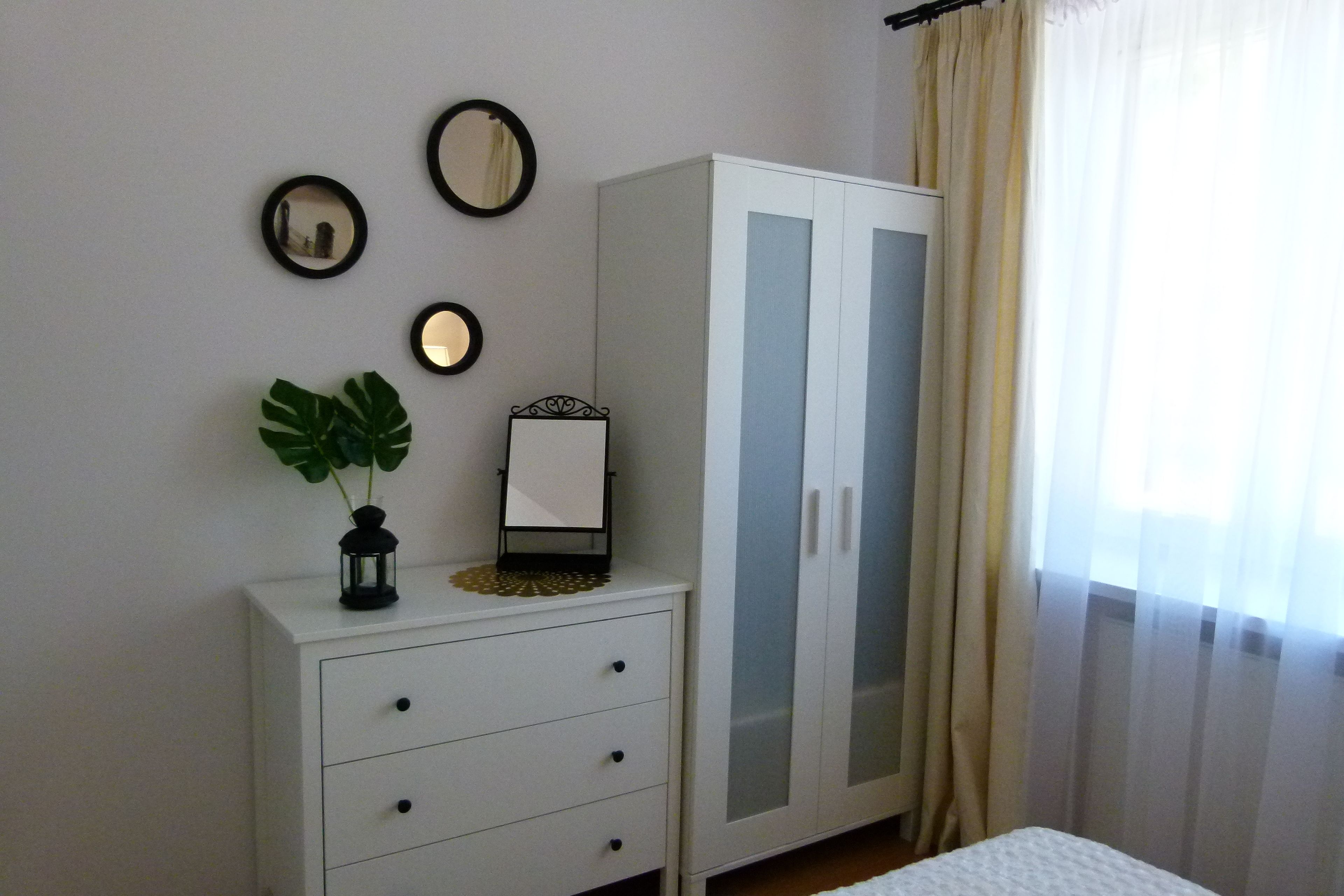 A bedroom with a white wardrobe, a bedside table with a plant, and three wall mirrors.