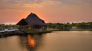 A thatched-roof structure by a calm body of water, likely a lodge or pavilion, with a bridge leading to it. - Stephen Margolis Resort (Harare)