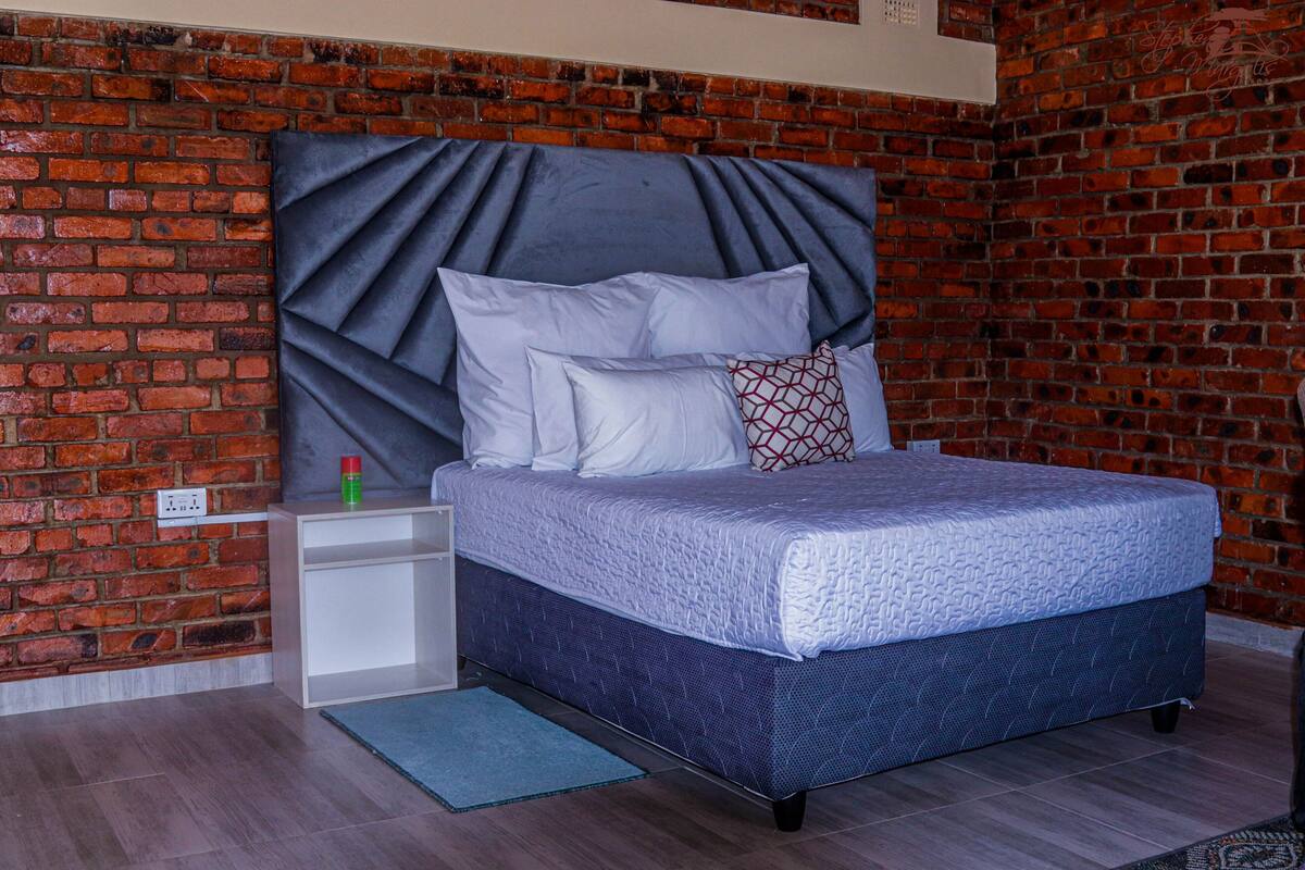 Ein Schlafzimmer mit Backsteinwand, einem Bett mit blauer Kopfteil, weißer Bettwäsche und einem kleinen Nachttisch mit einer roten Flasche.