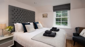 A bedroom with a large tufted headboard, a neatly made bed with multiple pillows, a bedside table with a lamp and flowers, and a window with a view of greenery. - Mill House (Crieff)