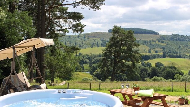 A hot tub with a swing chair and picnic table, set against a scenic hillside view. - Brae Cottage (Crieff)