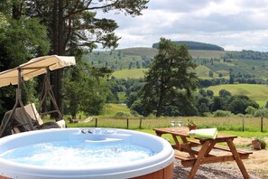 A hot tub with a swing chair and picnic table, set against a scenic hillside view. - Brae Cottage (Crieff)