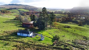A rural landscape with rolling hills, a white house with a red roof, and a large tree. - Brae Cottage (Crieff)